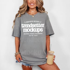 May include: A gray t-shirt with the text "trendsetter mockups" in white. The shirt is worn with denim shorts and accessories. A glass with a straw and a beverage is held in the hand.