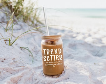 Op de afbeelding: Een heldere glazen beker met een bamboe deksel en een helder rietje, gevuld met een bruine drank. De beker heeft de woorden "TREND SETTER" in witte letters. De achtergrond is een zandstrand met de zee in de verte.