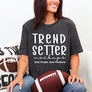Puede incluir: Camiseta gris oscuro con el texto "TREND SETTER mockups" en blanco. Se sostiene un balón de fútbol americano marrón y blanco en el regazo. Un cojín con un diseño de fútbol americano está en el fondo.