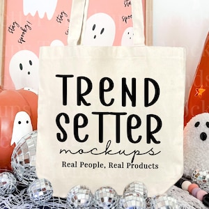Puede incluir: Un bolso tote color crema con el texto "TREND SETTER mockups Real People, Real Products" en negro. Está rodeado de decoraciones de Halloween, incluyendo una calabaza, un fantasma y bolas de discoteca.