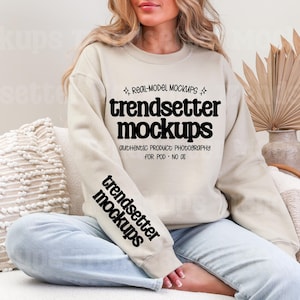 Könnte beinhalten: Ein beiges Sweatshirt mit dem Text "trendsetter mockups" in Schwarz auf der Vorderseite und am Ärmel. Das Sweatshirt wird von einer Person getragen. Das Bild enthält ein dekoratives Kissen und getrocknete Pflanzen.