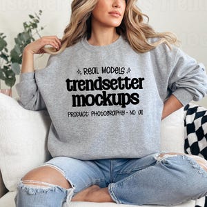 Op de afbeelding: Een grijs gemêleerd sweatshirt met ronde hals en zwarte tekst REAL MODELS trendsetter mockups PRODUCT PHOTOGRAPHY NO AI met kleine steraccenten, gecombineerd met een lichtblauwe spijkerbroek met gerafelde gaten bij de knieën.
