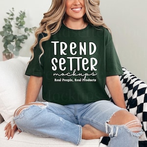 May include: Dark green t-shirt with the text "TREND SETTER mockups Real People, Real Products" in white. The shirt is paired with light blue ripped jeans. The person is sitting on a white couch with a black and white checkered blanket.