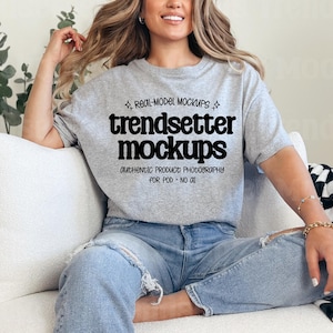 Peut inclure: T-shirt gris chiné avec l'inscription "trendsetter mockups" en noir. Le t-shirt porte également la mention "authentic product photography for pod - no oil". Le modèle porte un jean bleu et est assis sur un canapé blanc.