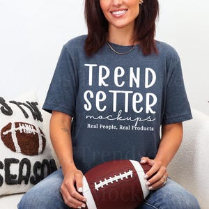 Puede incluir: Una camiseta azul marino con el texto blanco "Trend Setter mockups Real People, Real Products". La persona sostiene un balón de fútbol americano marrón. Se ve un cojín con un diseño de balón de fútbol.