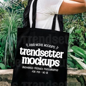 May include: Black tote bag with white text that reads "trendsetter mockups, authentic product photography for POD - NO AI." The bag has black handles and is being carried outdoors. The background is blurred with green foliage.