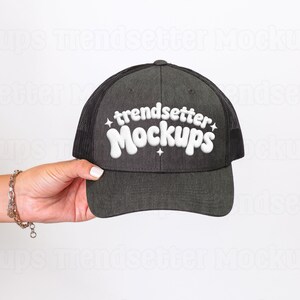 以下が含まれることがあります： 黒いメッシュのバックが付いたチャコールグレーのトラッカーハット。「trendsetter Mockups」の文字が白いスタイリッシュな文字で表示されています。帽子は人が持っています。