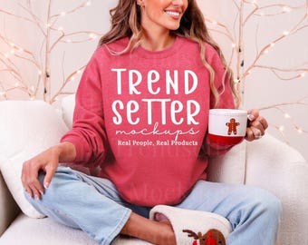 May include: A red crewneck sweatshirt featuring the white text TREND SETTER mockups Real People, Real Products. The sweatshirt is paired with light blue denim jeans and a white mug with a gingerbread man graphic.