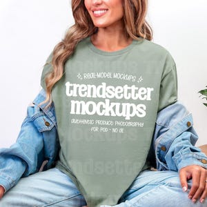 May include: Sage green t-shirt with the text "trendsetter mockups" in a bold, white font. The shirt is worn with a light blue denim jacket and light wash jeans. A small potted plant is visible in the background.