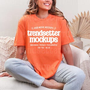 May include: An orange t-shirt with the text "trendsetter mockups" in white. The shirt is worn by a person sitting on a cream-colored chair. The background includes a decorative dried plant arrangement and a patterned pillow.