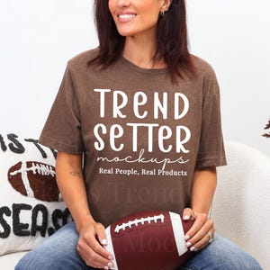 Puede incluir: Una camiseta marrón con el texto "TREND SETTER mockups Real People, Real Products" en blanco. Una persona sostiene un balón de fútbol americano marrón y blanco. Una almohada blanca con un diseño de fútbol americano está en el fondo.