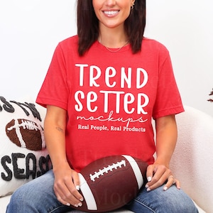 Puede incluir: Camiseta roja con el texto "TREND SETTER mockups Real People, Real Products". Un balón de fútbol americano marrón y blanco se sostiene en el regazo. Se ve una almohada con un diseño de fútbol y las palabras "THIS SEASON".