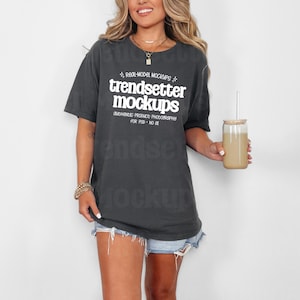 Könnte beinhalten: Ein anthrazitfarbenes T-Shirt mit dem weißen Text "trendsetter mockups". Das Shirt wird mit Jeansshorts kombiniert. Ein klares Glas mit einem beigefarbenen Getränk und einem Strohhalm wird in der Hand gehalten.