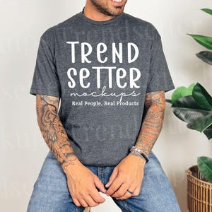 May include: A dark gray t-shirt with the text "TREND SETTER mockups Real People, Real Products" in white. The person is wearing blue jeans and sitting on a wooden stool. The person has tattoos on their arms.