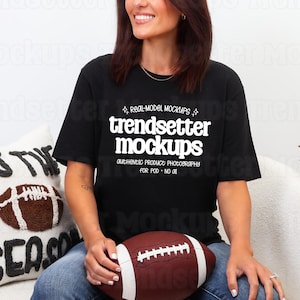 May include: A black short-sleeve t-shirt featuring white text that reads trendsetter mockups, real-model mockups, and authentic product photography for pod - no ai. The shirt is worn with blue jeans and held with a brown and white football.