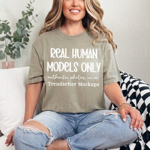 May include: An olive green t-shirt with the text "REAL HUMAN MODELS ONLY" in white. The shirt is paired with blue jeans. The model is sitting on a white couch with a black and white checkered blanket.