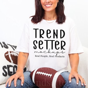 Puede incluir: Una camiseta blanca con el texto "Trend Setter mockups Real People, Real Products" en negro. Un balón de fútbol americano marrón y blanco se sostiene en el regazo. Una almohada blanca con un diseño de fútbol americano está en el fondo.