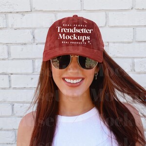 Puede incluir: Una gorra de béisbol granate con el texto "REAL PEOPLE Trendsetter Mockups REAL PRODUCTS" en blanco. La gorra se usa con gafas de sol con montura dorada.