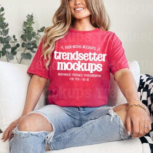 May include: A red t-shirt with the text "trendsetter mockups" in white. The shirt is worn with ripped blue jeans. The model is sitting on a white couch with a black and white checkered blanket.