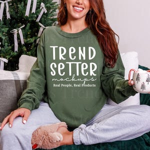 Op de afbeelding: Een olijfgroene sweatshirt met de tekst "TREND SETTER mockups Real People, Real Products" in het wit. De persoon houdt een witte mok vast met een zwart boomontwerp en een rood handvat. De persoon draagt een grijze joggingbroek en fluffy pantoffels.