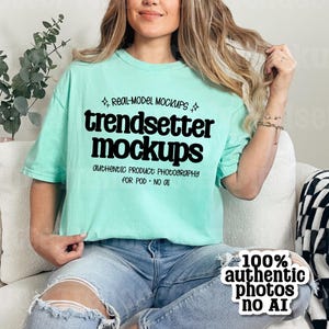May include: A light green t-shirt with the text "trendsetter mockups" in bold black letters. The shirt also has the text "authentic product photography for POD - NO AI". The model is wearing blue jeans and sitting on a white couch.