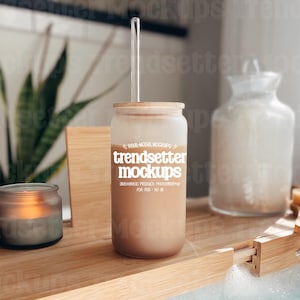 May include: A frosted glass tumbler with a bamboo lid and a clear straw, filled with a light brown liquid. The tumbler has the text "trendsetter mockups" printed on it. A lit candle in a glass jar and a decorative jar are also visible.