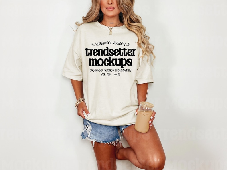 Front view mockup of a real person model wearing an oversized Comfort Colors 1717 t-shirt in Ivory. The model is wearing denim shorts and holding a glass of iced coffee, standing in a bright, minimalist setting.