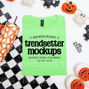 May include: A lime green t-shirt with the text "trendsetter mockups" is surrounded by Halloween-themed items. An orange tumbler with white ghost designs, a black and white checkered blanket, and pumpkin and bat decorations are visible.