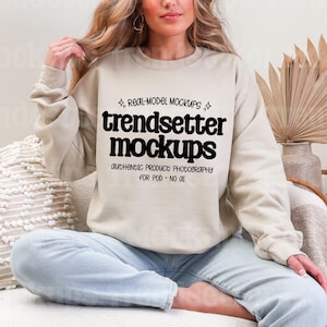 May include: A beige sweatshirt with the text "trendsetter mockups" in black. The sweatshirt is worn by a person sitting on a white couch. The image includes dried plants in a vase.