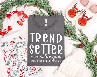 May include: A dark gray t-shirt with white text that reads TREND SETTER mockups Real People, Real Products, surrounded by holiday decorations including pine branches, red and white candy ornaments, reindeer slippers, and white pajamas.