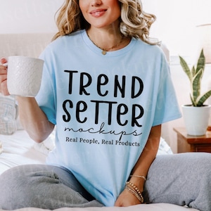 Puede incluir: Una camiseta azul claro con la frase "TREND SETTER mockups" en negro. La camiseta se combina con un pantalón de chándal gris. Se sostiene una taza blanca. El texto "Real People, Real Products" está debajo.