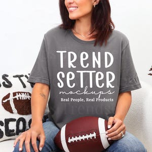Puede incluir: Una camiseta gris con el texto "Trend Setter mockups Real People, Real Products" en blanco. Un balón de fútbol americano marrón y blanco se sostiene en el regazo. Un cojín decorativo con un diseño de fútbol está a la izquierda.