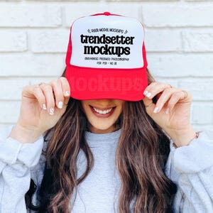 Könnte beinhalten: Eine rot-weiße Trucker-Mütze mit dem Text "trendsetter mockups" in Schwarz. Die Mütze wird von einer Person hochgehalten. Der Hintergrund ist eine weiße Backsteinmauer.
