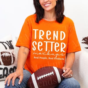 Puede incluir: Una camiseta naranja con las palabras "TREND SETTER mockups" en blanco. Un balón de fútbol marrón y blanco se sostiene en el regazo. Un cojín decorativo con un diseño de fútbol está a la izquierda.