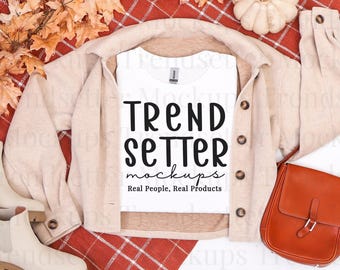 May include: A white t-shirt featuring the black text TREND SETTER mockups Real People, Real Products, layered under a beige button-down shirt, accompanied by a brown leather shoulder bag and cream-colored ankle boots.