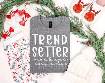 May include: A heather gray crew neck t-shirt featuring the white text TREND SETTER mockups Real People, Real Products, accompanied by white and red patterned pajama pants, white reindeer slippers, and a white knit pom-pom beanie.