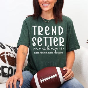 Puede incluir: Una camiseta verde oscuro con el texto "TREND SETTER mockups Real People, Real Products" en blanco. Se sostiene un balón de fútbol marrón en el regazo. Una almohada con un diseño de fútbol está en el fondo.