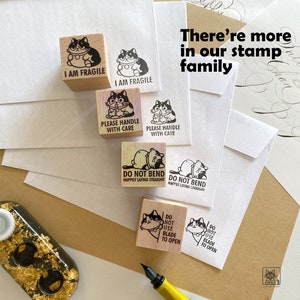 May include: Wooden stamps with cat illustrations and phrases like "I AM FRAGILE" and "PLEASE HANDLE WITH CARE." The stamps are arranged with stamped images on white envelopes. The text "There're more in our stamp family" is also visible.