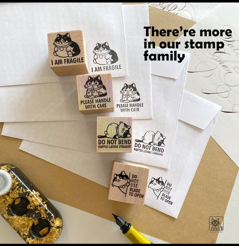 May include: Wooden stamps with cat illustrations and phrases like "I AM FRAGILE" and "PLEASE HANDLE WITH CARE." The stamps are arranged with stamped images on white envelopes. The text "There're more in our stamp family" is also visible.