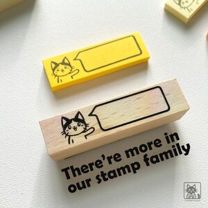 May include: Two rectangular stamps with a cat illustration and speech bubble design. One stamp is yellow, the other is wood-toned. Text on the wooden stamp reads "There're more in our stamp family."