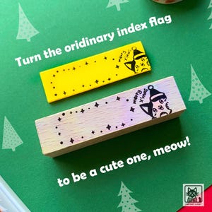 May include: Two wooden index flag stamps with a cat wearing a Santa hat and the text "merry x'mas". The stamps are on a green background with a small Christmas tree.