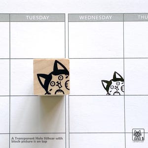 May include: A wooden stamp with a black and white cat face design, next to a printed cat image on a calendar grid. The stamp is square and the cat image is repeated on the calendar. Text at the bottom reads "A Transparent Holo Stikcer with black picture is on top."