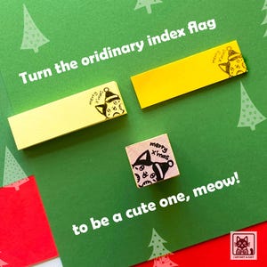 May include: Yellow index flags and a small wooden stamp with a cat illustration and the text "merry x'mas" on a green background with white Christmas tree designs. The text "Turn the ordinary index flag" and "to be a cute one, meow!" are also visible.