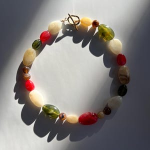 Könnte beinhalten: Eine farbenfrohe Halskette mit ovalen Perlen in Rot, Grün, Gelb, Braun und Creme. Die Perlen sind aufgereiht und mit einem goldfarbenen Knebelverschluss versehen. Ein einzigartiges Accessoire, perfekt, um jedem Outfit einen Farbtupfer zu verleihen.