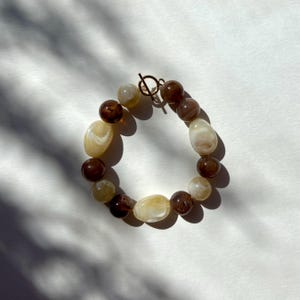 Può includere: Un braccialetto di perline marroni, crema e ambra. Il braccialetto ha una chiusura a levetta color oro. Le perline variano per dimensioni e forma, creando un aspetto unico. Il braccialetto è esposto su una superficie bianca.