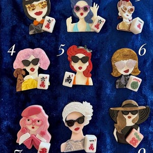 May include: A collection of nine acrylic brooches featuring stylized figures wearing various hats, hairstyles, and sunglasses, each holding a mahjong tile. The figures display diverse colors including pink, red, brown, and black, with detailed facial features and accessories.
