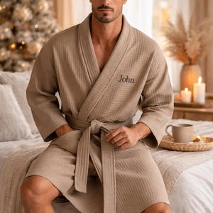 May include: A beige waffle-weave robe with a matching belt and pockets. The word "John" is embroidered on the chest. The robe is knee-length and worn open, with the model seated on a bed. The background includes a Christmas tree.
