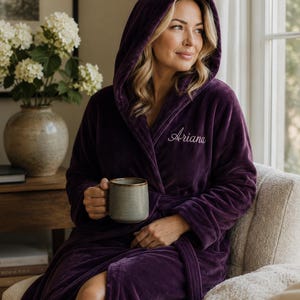 May include: A deep purple plush hooded robe featuring long sleeves, a tie waist, and the name Ariana embroidered in white script on the left chest. The robe is made of a soft, velvety fabric and includes a hood that rests over the shoulders.