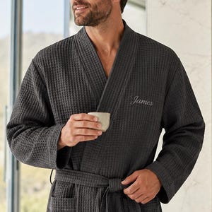 May include: Dark gray waffle-weave robe with a shawl collar and a matching belt. The robe has a pocket on the front and the name "James" embroidered on the chest. The robe is made of a soft, textured material.