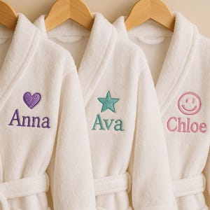 May include: Three white plush bathrobes hanging on wooden hangers. Each robe has a different embroidered design and name: a purple heart with "Anna", a teal star with "Ava", and a pink smiley face with "Chloe". The robes have matching belts and pockets.